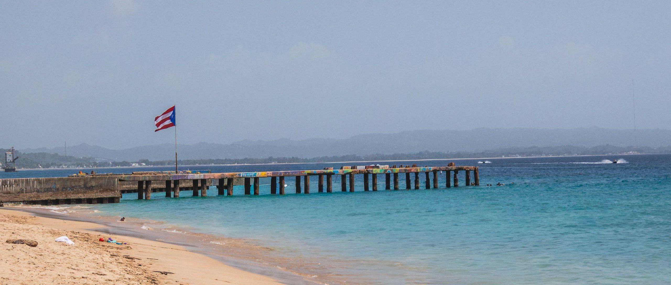 Calm beach view in Aguadilla, Puerto Rico with pier and national flag waving gently.